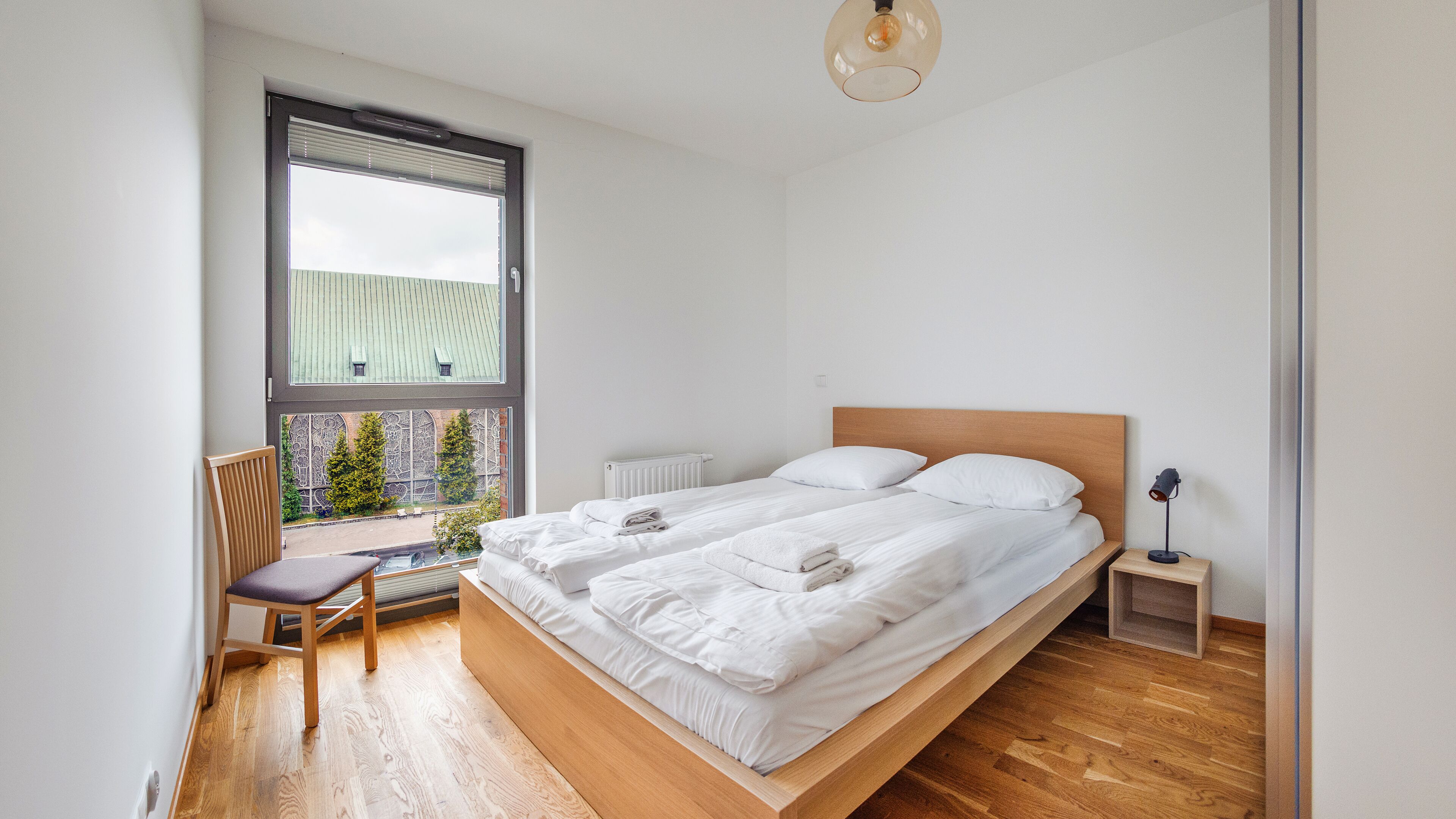A bedroom with a wooden bed, a chair, a desk, and a window with a view of trees and a building.