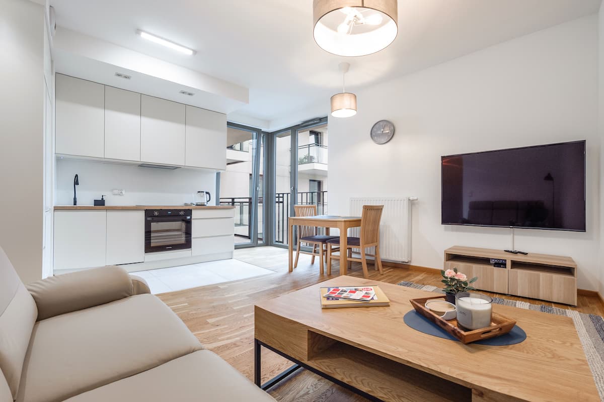 Un apartamento moderno con sala de estar, comedor y cocina. Cuenta con un televisor de pantalla plana, un sofá, una mesa de centro con revistas y una vela, y vistas al balcón.