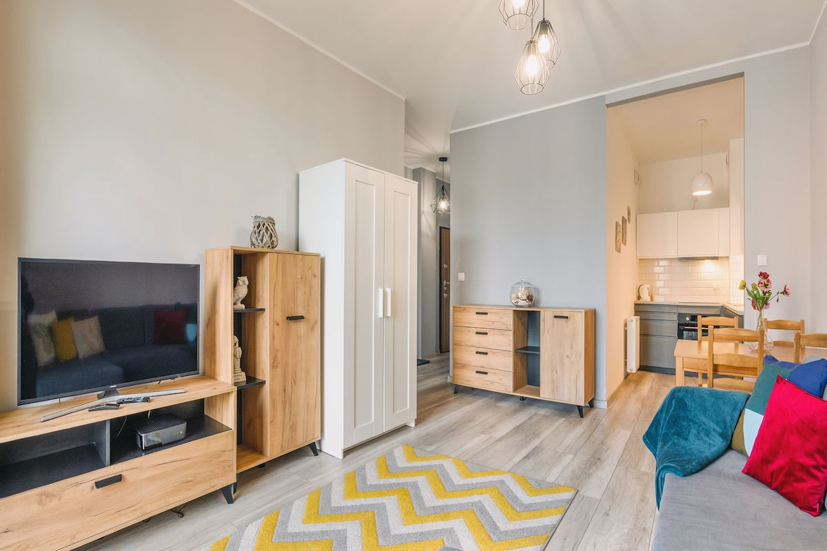 Una sala de estar moderna con televisor, armarios de madera, un sofá con cojines de colores y una cocina al fondo.
