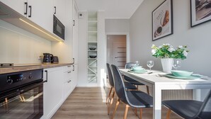 Een moderne keuken met een ingebouwde oven, een eethoek met een witte tafel gedekt voor twee personen, en een witte vaas met bloemen aan de muur.