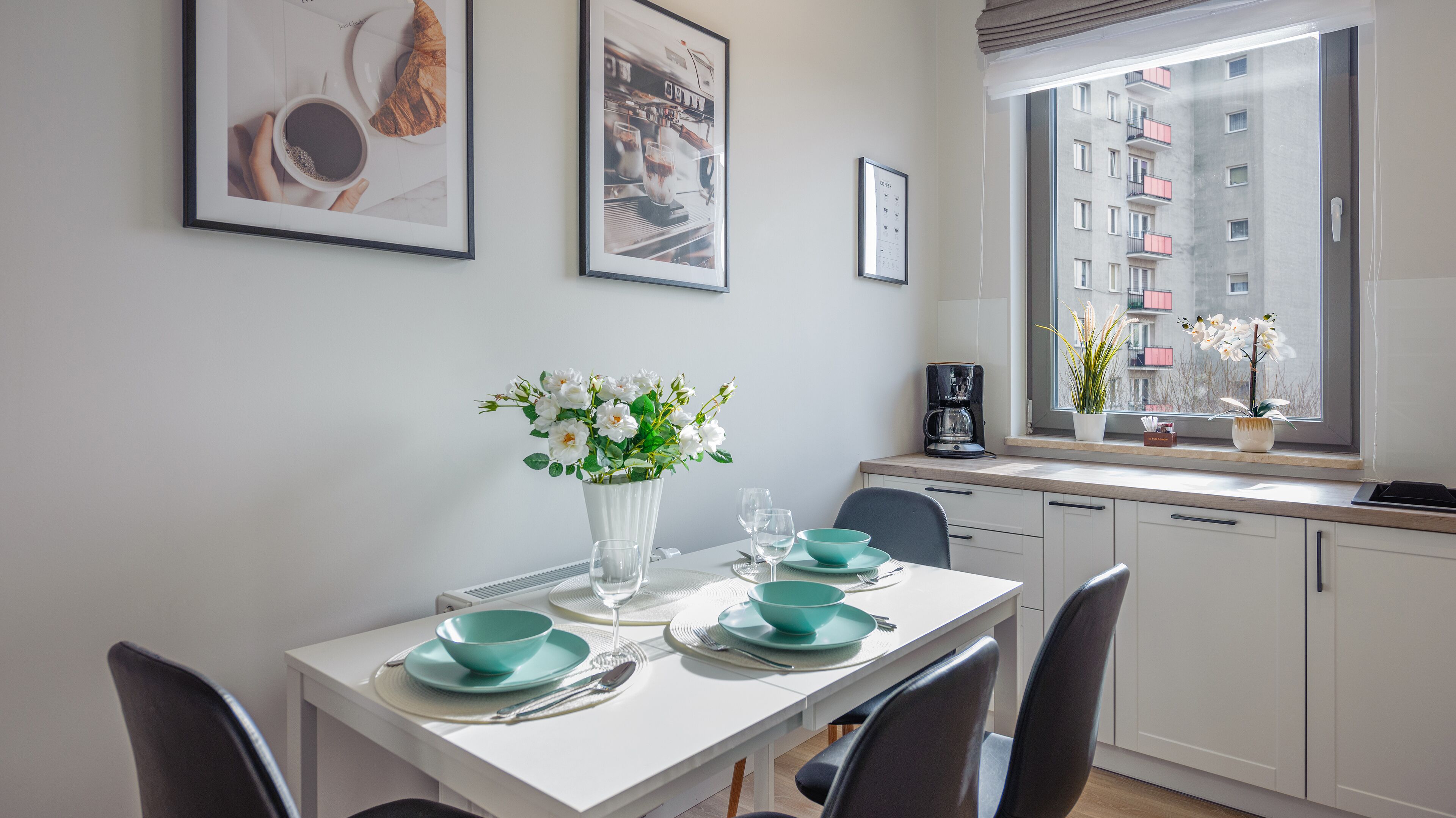 A modern kitchen with white cabinets, a dining table set with teal plates and glasses, a coffee machine, and a window with a view of a building.