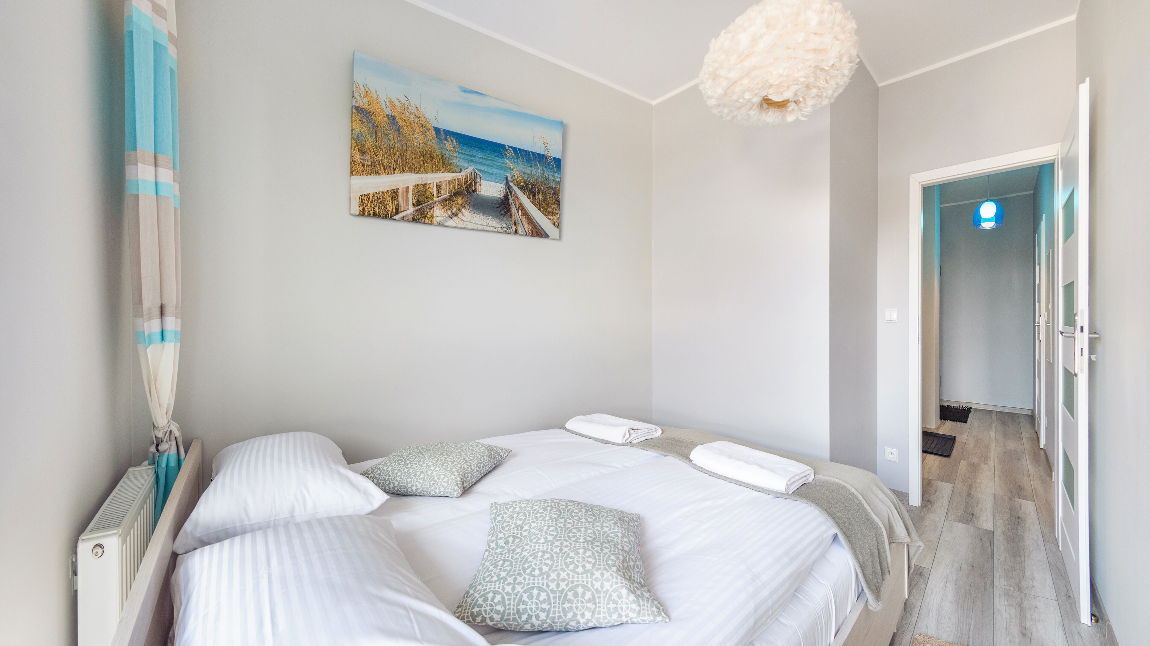 A bedroom with a bed, pillows, a painting of a beach, and a hallway leading to a door.