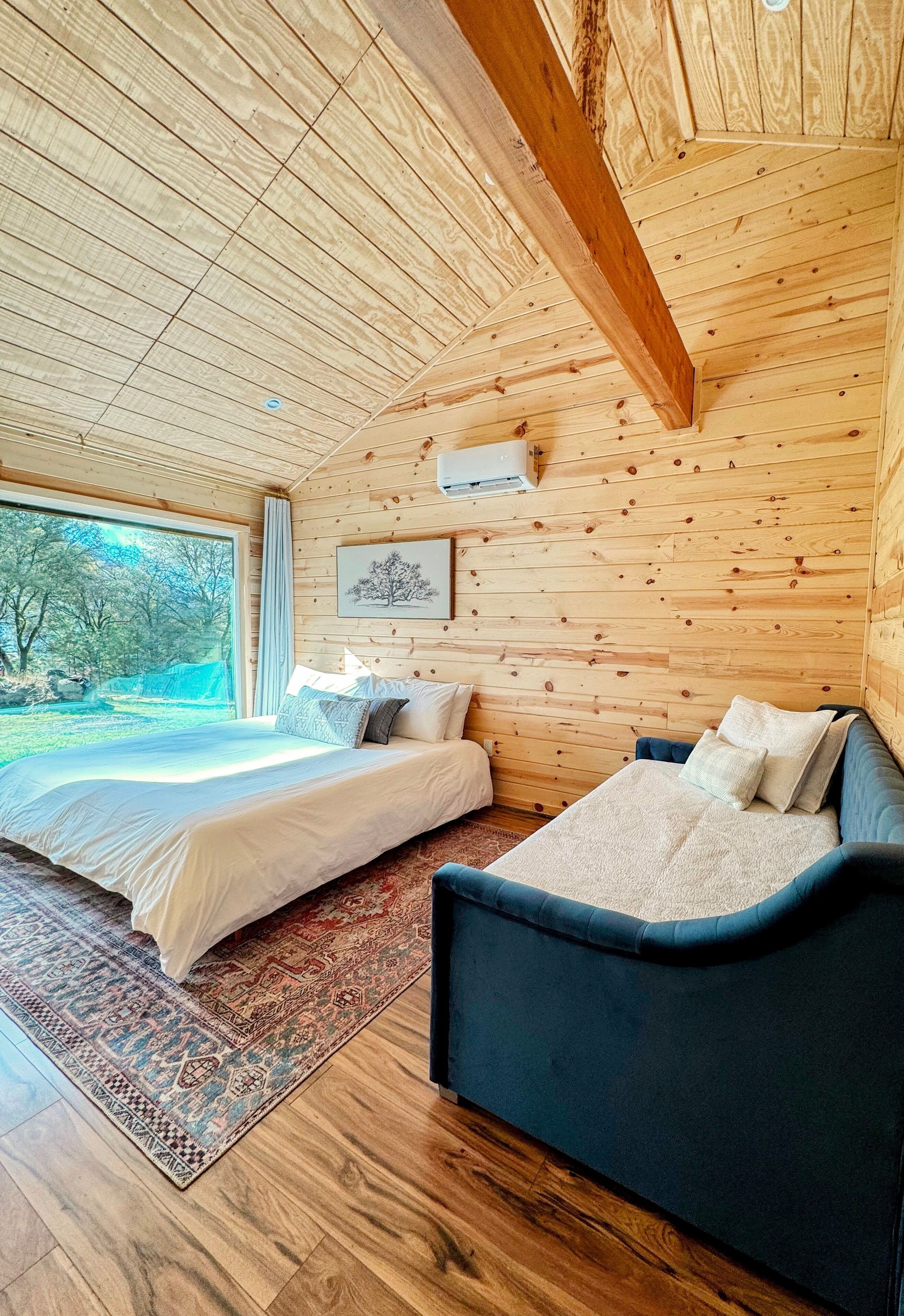 A wooden cabin bedroom with two beds, a large window, and a framed picture on the wall.