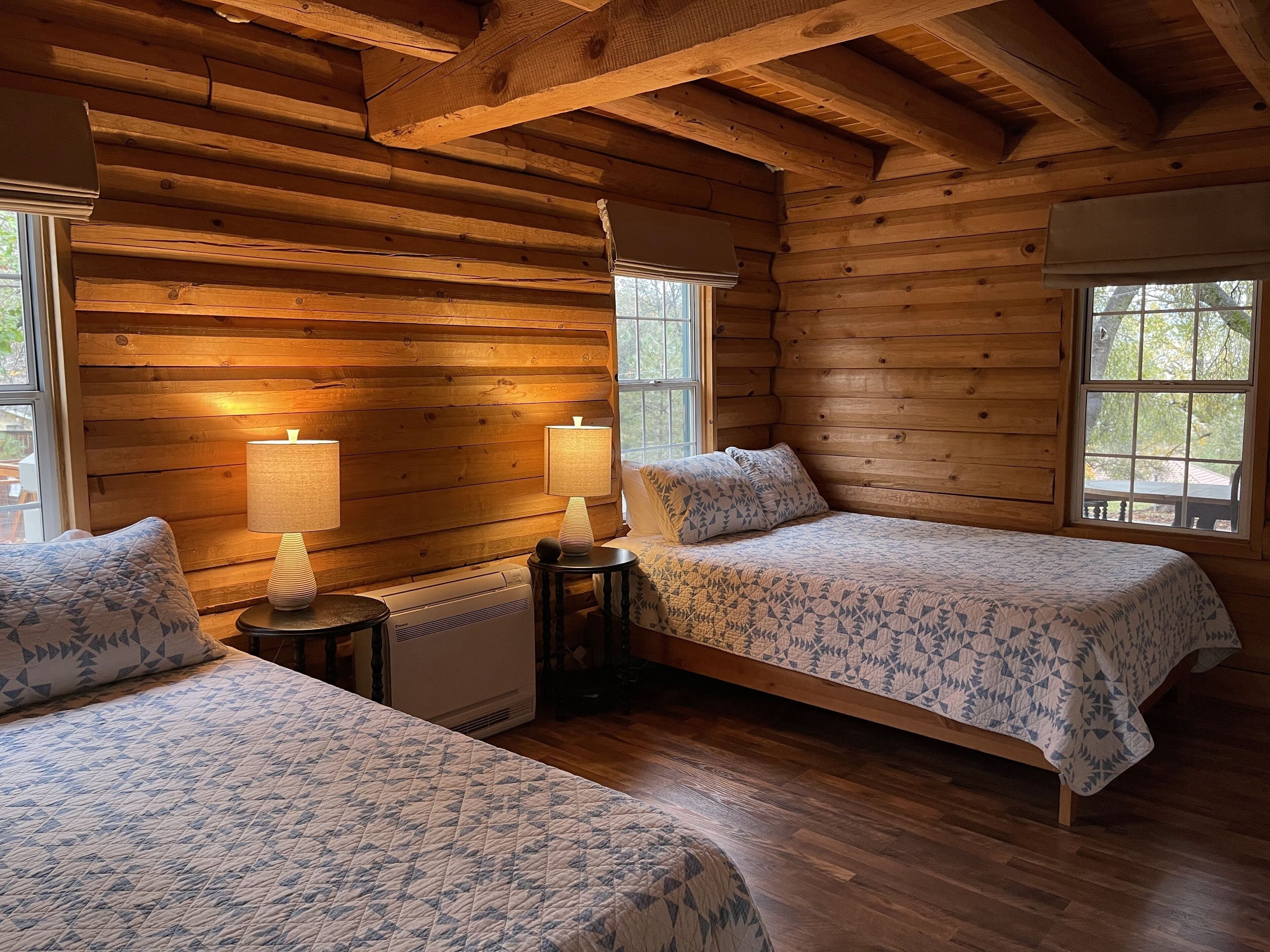 A log cabin bedroom with two beds, each with a patterned quilt, bedside lamps, and a heater.