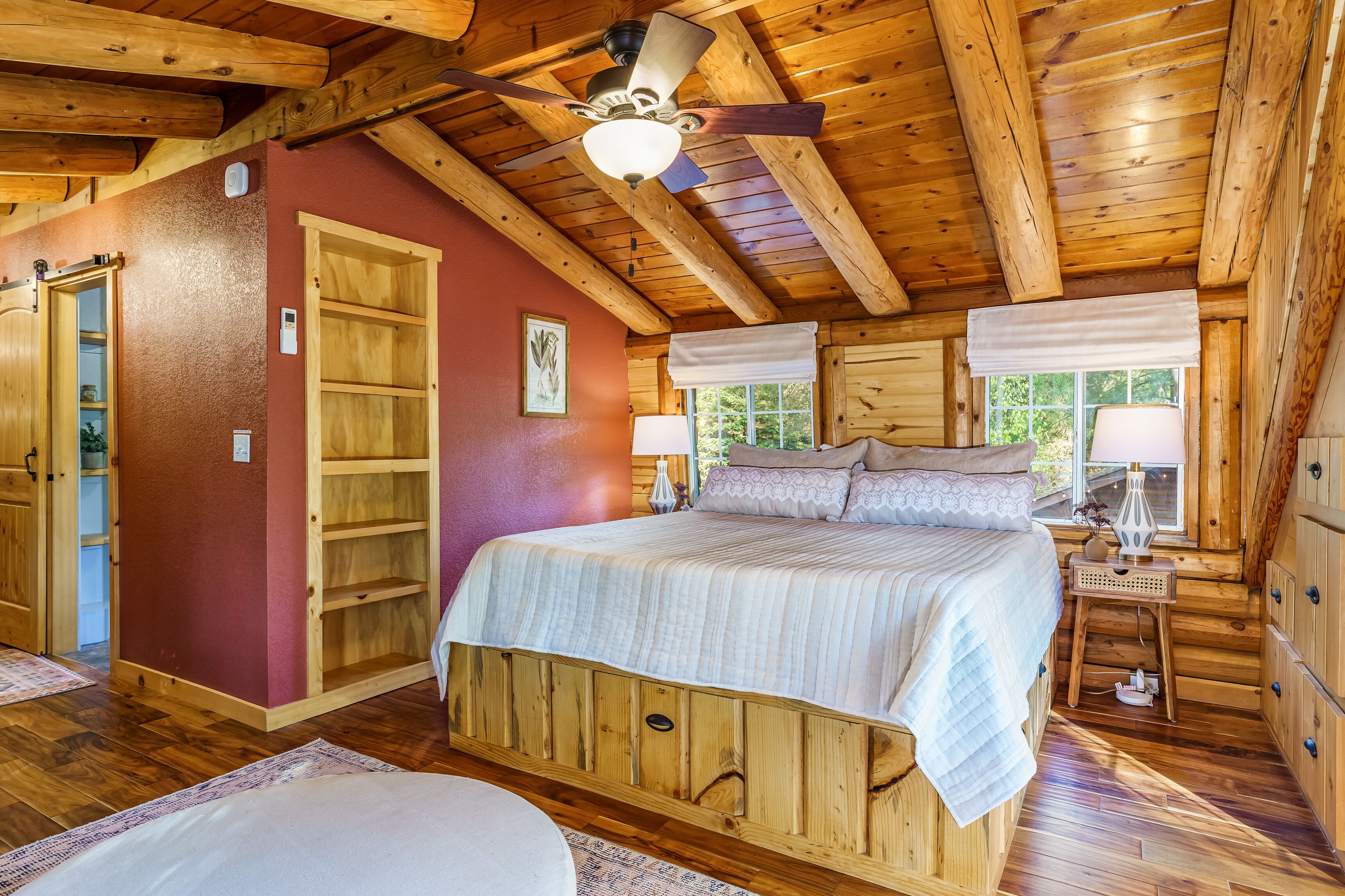 A wooden cabin bedroom with a bed, a ceiling fan, a window with a view, and a wooden wardrobe.