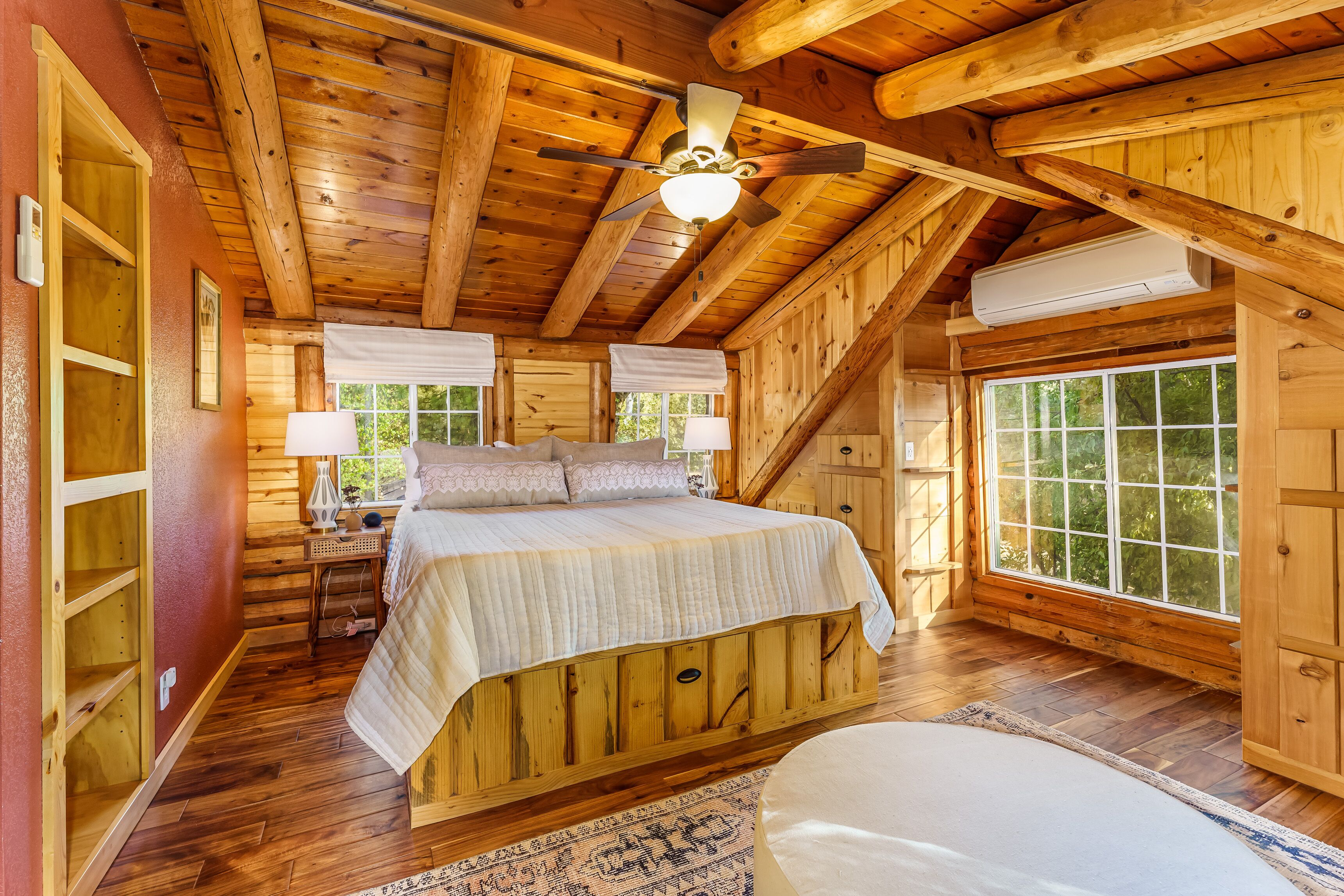 A wooden cabin bedroom with a bed, two windows, a ceiling fan, and an air conditioning unit.
