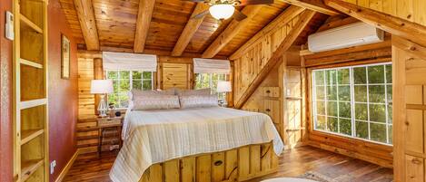 A wooden cabin bedroom with a bed, two windows, a ceiling fan, and an air conditioning unit.