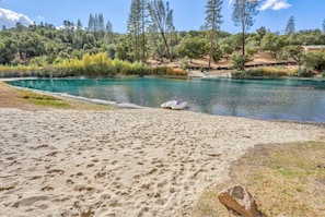 A sandy beach area with a clear blue pool, surrounded by trees and a hilly landscape. - Oak Pond Retreat Stocked pond/pool table/BBQ Grill/EV Charger (Oakhurst)