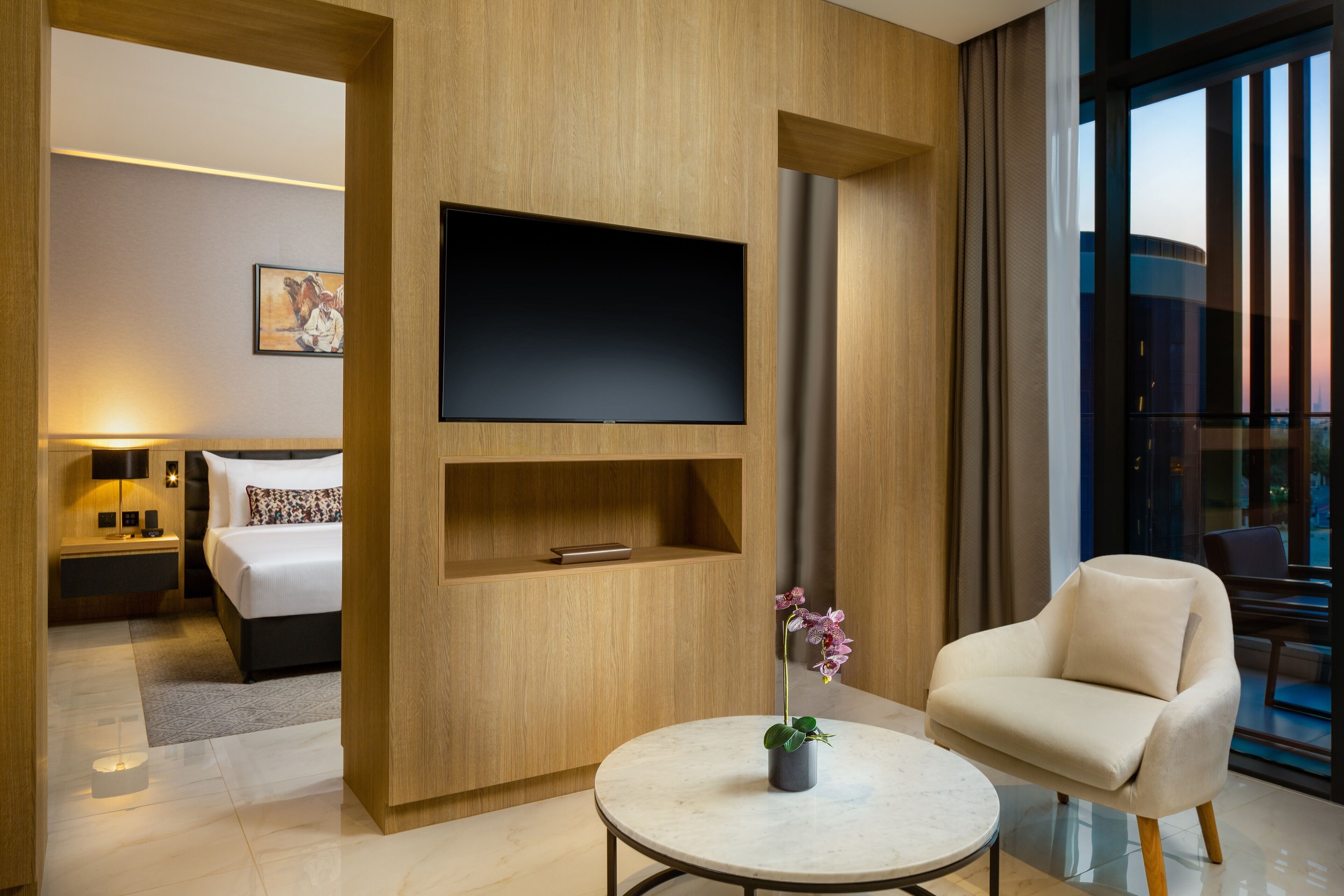A modern hotel room with a large flat-screen TV mounted on a wooden wall, a comfortable sofa, a round coffee table with a potted orchid, a neatly made bed, and a large window offering a city view.