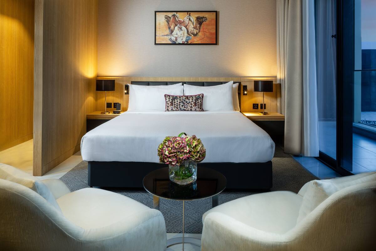 Ett hotellrum med en stor säng, två sängbord med lampor, ett litet runt bord med en vas med blommor och två fåtöljer.