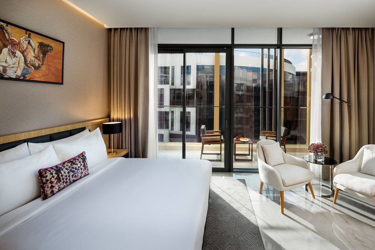 Ett hotellrum med en stor säng, en sittgrupp med två stolar, ett litet bord och ett stort fönster med utsikt över byggnader.