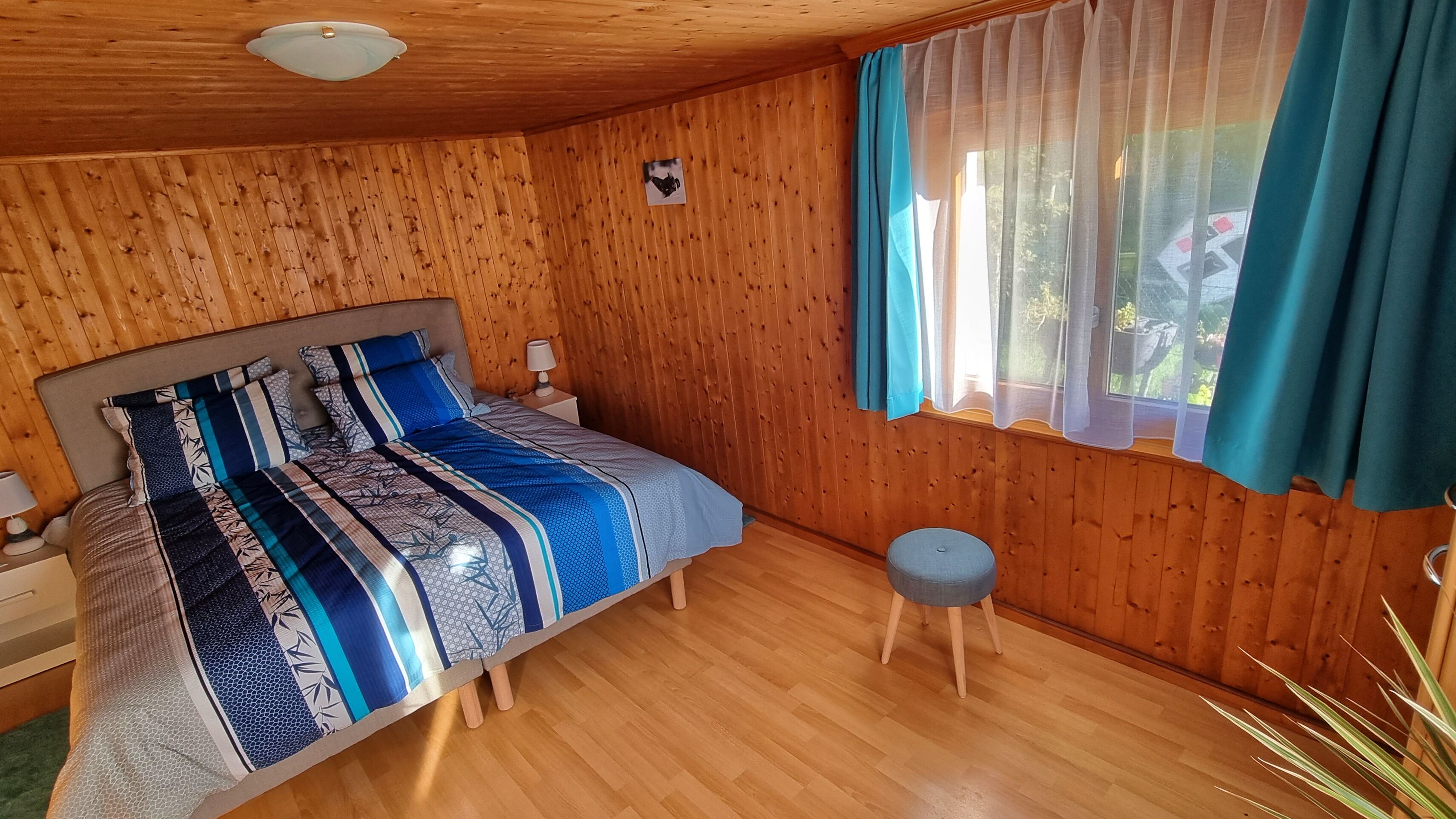 Een kamer met houten lambrisering, een bed, een klein krukje en een raam met gordijnen.