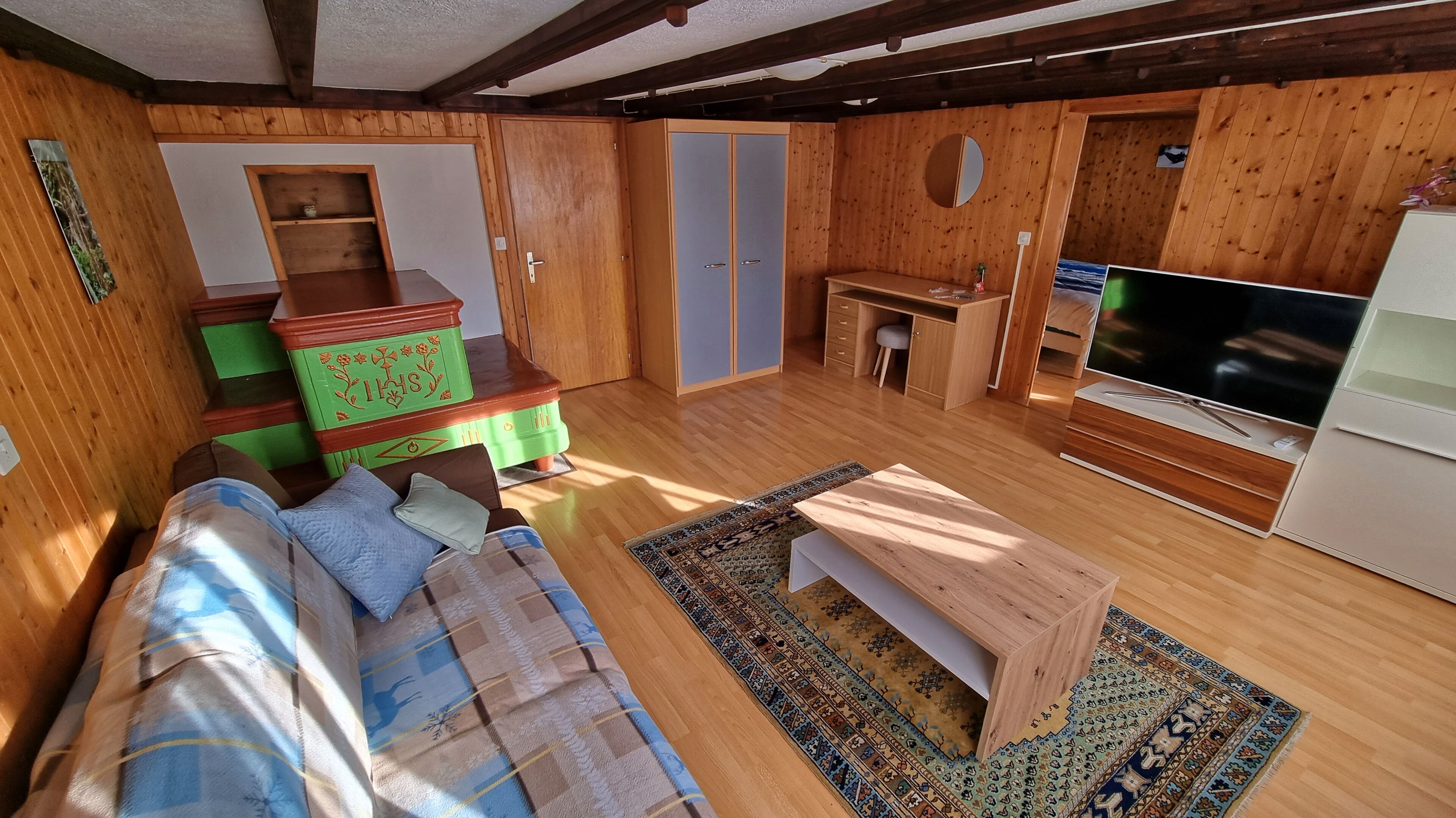 A compact room with a wooden floor, a sofa with patterned cushions, a small wooden table, a TV, and a built-in wardrobe.