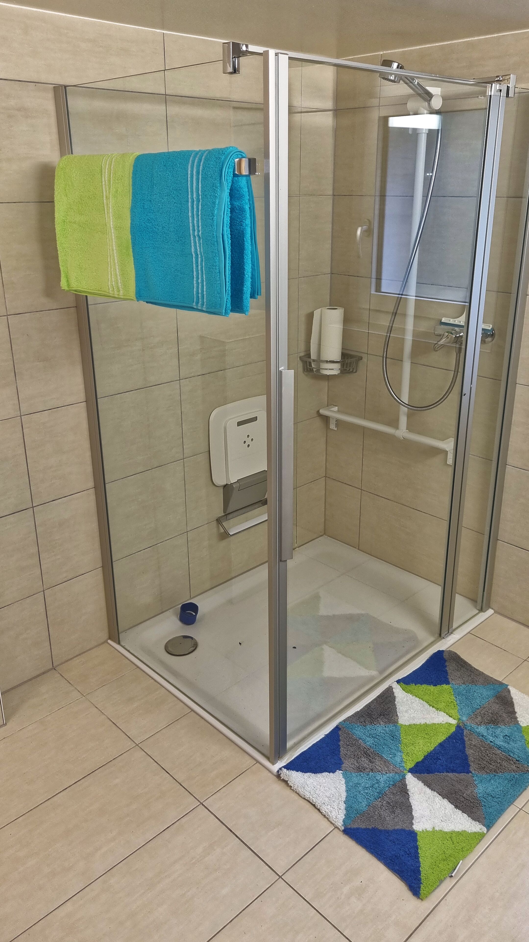 A glass-enclosed shower stall with a towel rack holding two towels, a hand dryer, and a paper towel dispenser.