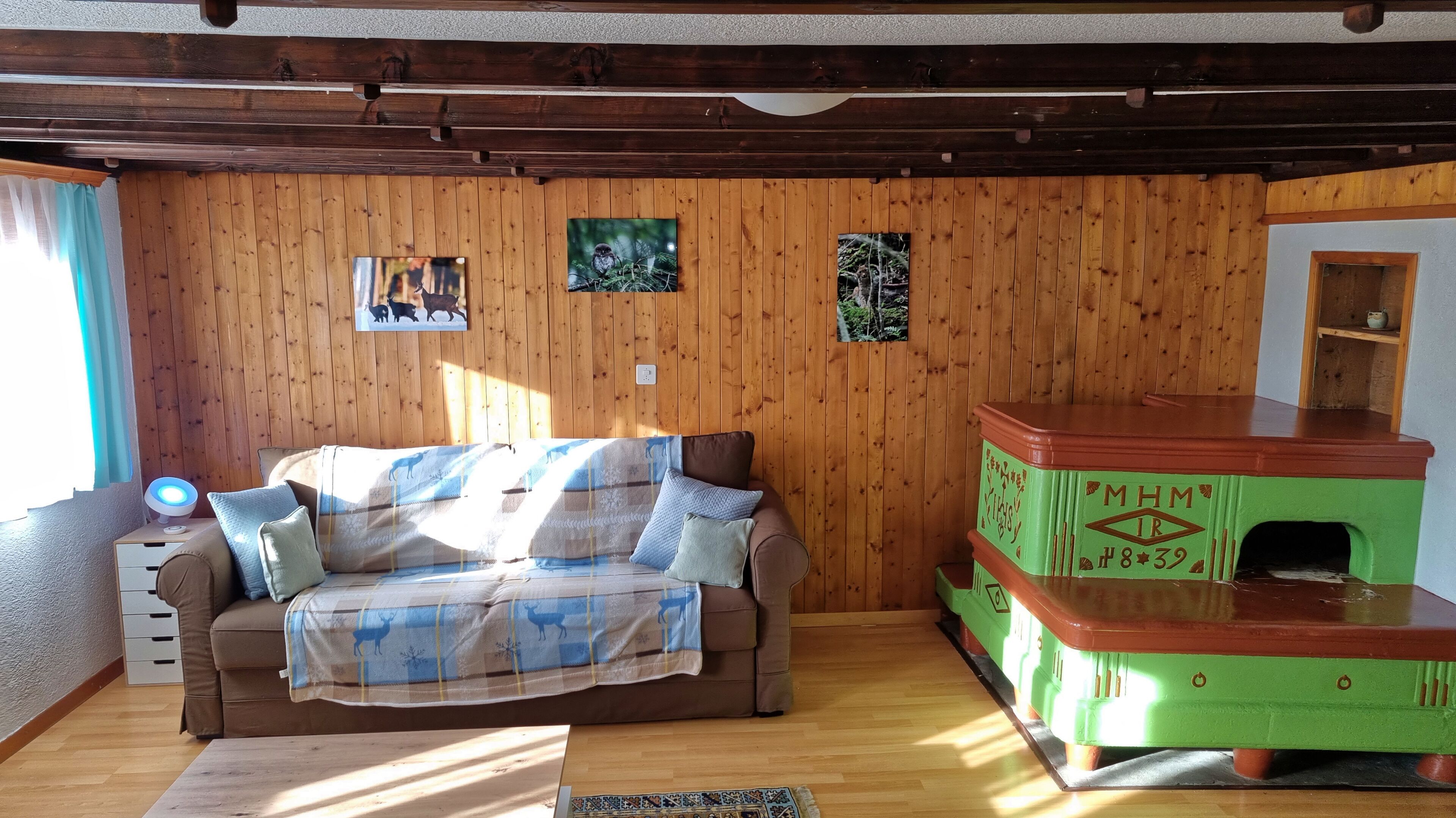 Une pièce chaleureuse avec un mur en bois, un canapé marron recouvert d’une couverture, un radiateur vert et rouge, et une petite étagère.