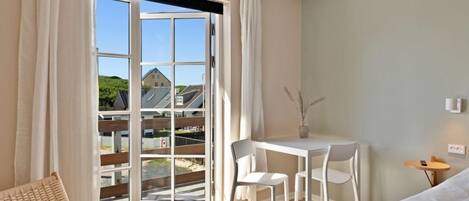 A hotel room with a large sliding glass door to a balcony, a small white table with two chairs, a wicker chair, and a bed with white bedding.
