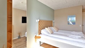 Un dormitorio moderno con cabecera de madera, cama con ropa de cama blanca, una mesita de noche pequeña y un televisor montado en la pared.