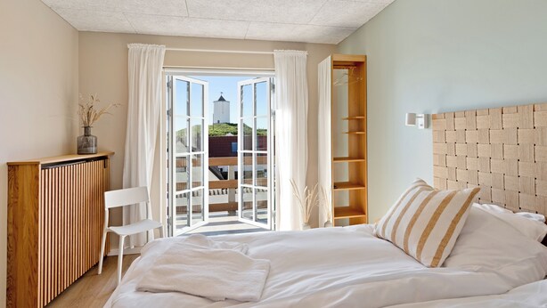Un dormitorio con cama, mesita de noche de madera, una silla, armario y vista a través de una puerta abierta.