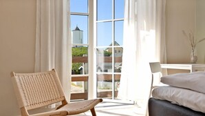 Una habitación con una silla de mimbre, una cama con ropa blanca, una mesita con un florero y vistas a un edificio a través de amplios ventanales.
