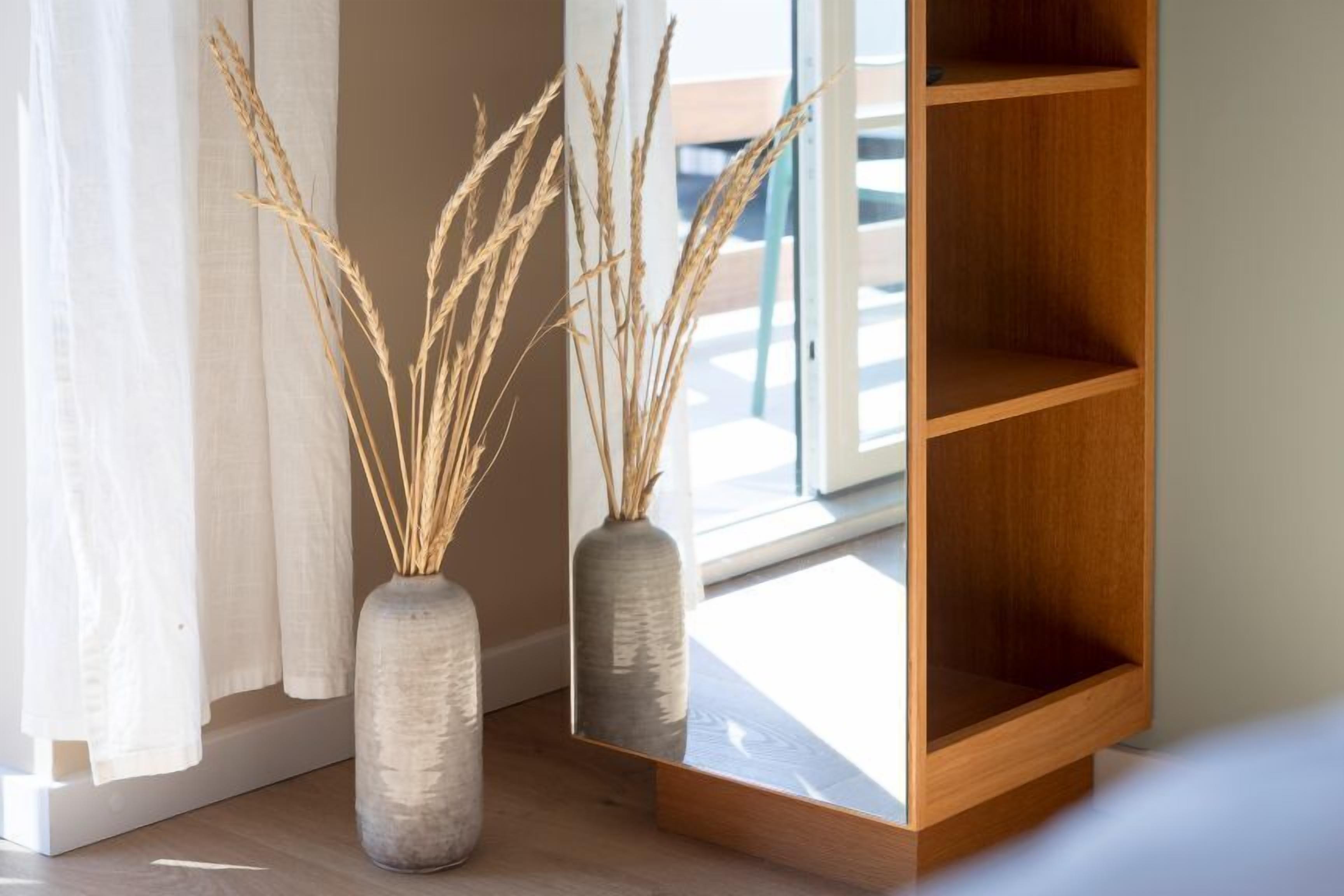 Two vases with dried plants on a wooden shelf near a window.