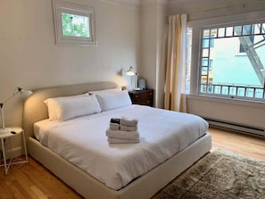 Un letto rifatto con un copriletto bianco e cuscini, una lampada da comodino, un tavolino con una lampada e vista all'esterno attraverso una finestra con tende.