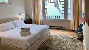 Habitación de hotel con una cama grande, una ventana con vistas a un edificio, una mesita de noche con lámpara y una obra de arte colgada en la pared.