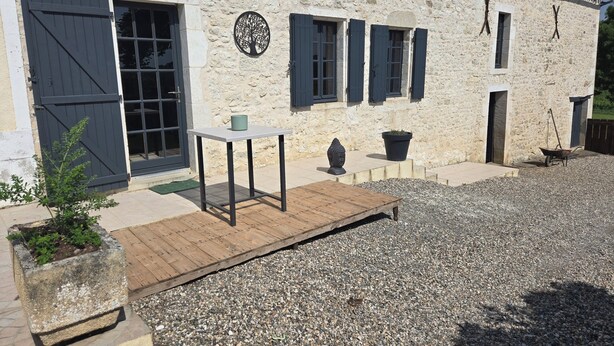 A stone building with a wooden deck, a wheelbarrow, and a potted plant. - Gîte les trois pierres (Riocaud)