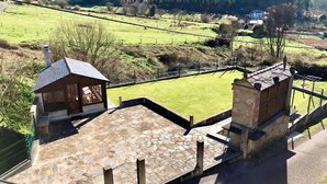 Un patio de piedra con una pequeña estructura de madera, rodeado por un jardín vallado y vistas al paisaje rural.
