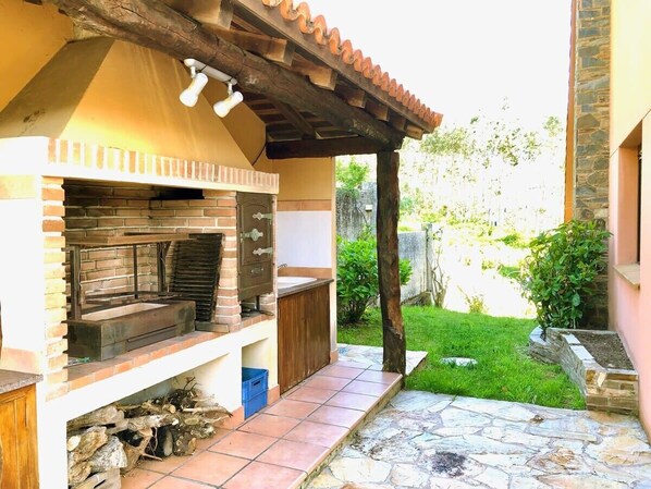 Eine Außenküche mit Backofen aus Ziegeln, Holzregalen und einem gepflasterten Weg, der zu einer überdachten Terrasse führt.