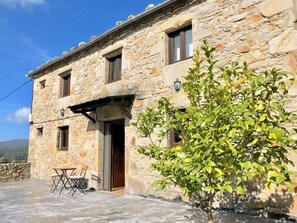A stone building with a wooden door, a small patio with a table and chairs, and a tree in front. - The Cliffs - Cottage Xilloi Beach (O Vicedo)