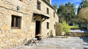 A stone building with a patio area, a small table with chairs, and a tree. - The Cliffs - Cottage Xilloi Beach (O Vicedo)