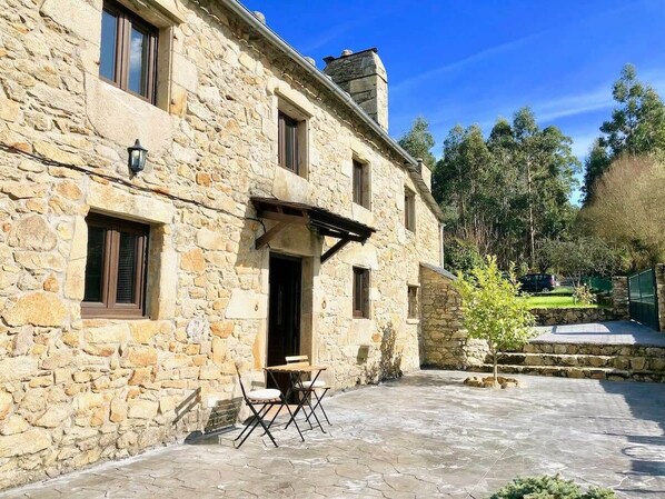 A stone building with a patio area, a small table with chairs, and a tree. - The Cliffs - Cottage Xilloi Beach (O Vicedo)