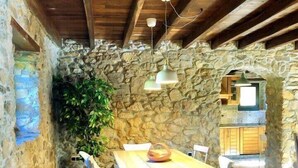 A rustic stone-walled dining area with a wooden table and chairs, stone archway, and a hanging light fixture. - The Cliffs - Cottage Xilloi Beach (O Vicedo)