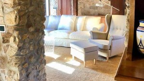 A cozy living room with a stone archway, wooden ceiling beams, a white sofa, a chair, and a small table. - The Cliffs - Cottage Xilloi Beach (O Vicedo)