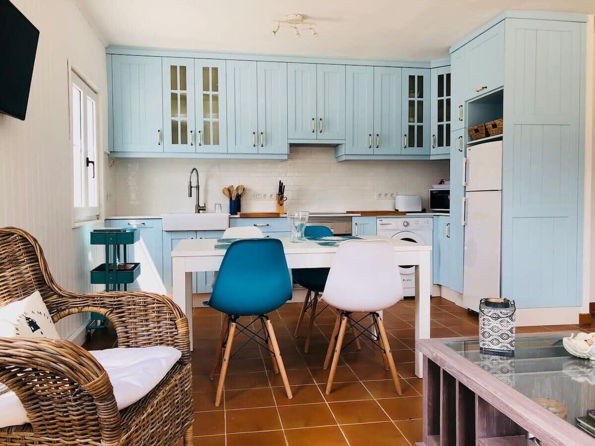 Een compacte keuken met lichtblauwe kastjes, een eettafel met twee stoelen en een rieten stoel met kussens.