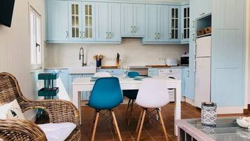 Een compacte keuken met lichtblauwe kastjes, een eettafel met twee stoelen en een rieten stoel met kussens.