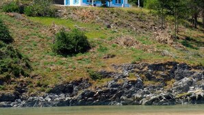 A coastal house with a white exterior and blue accents, situated on a grassy hill overlooking a sandy beach.