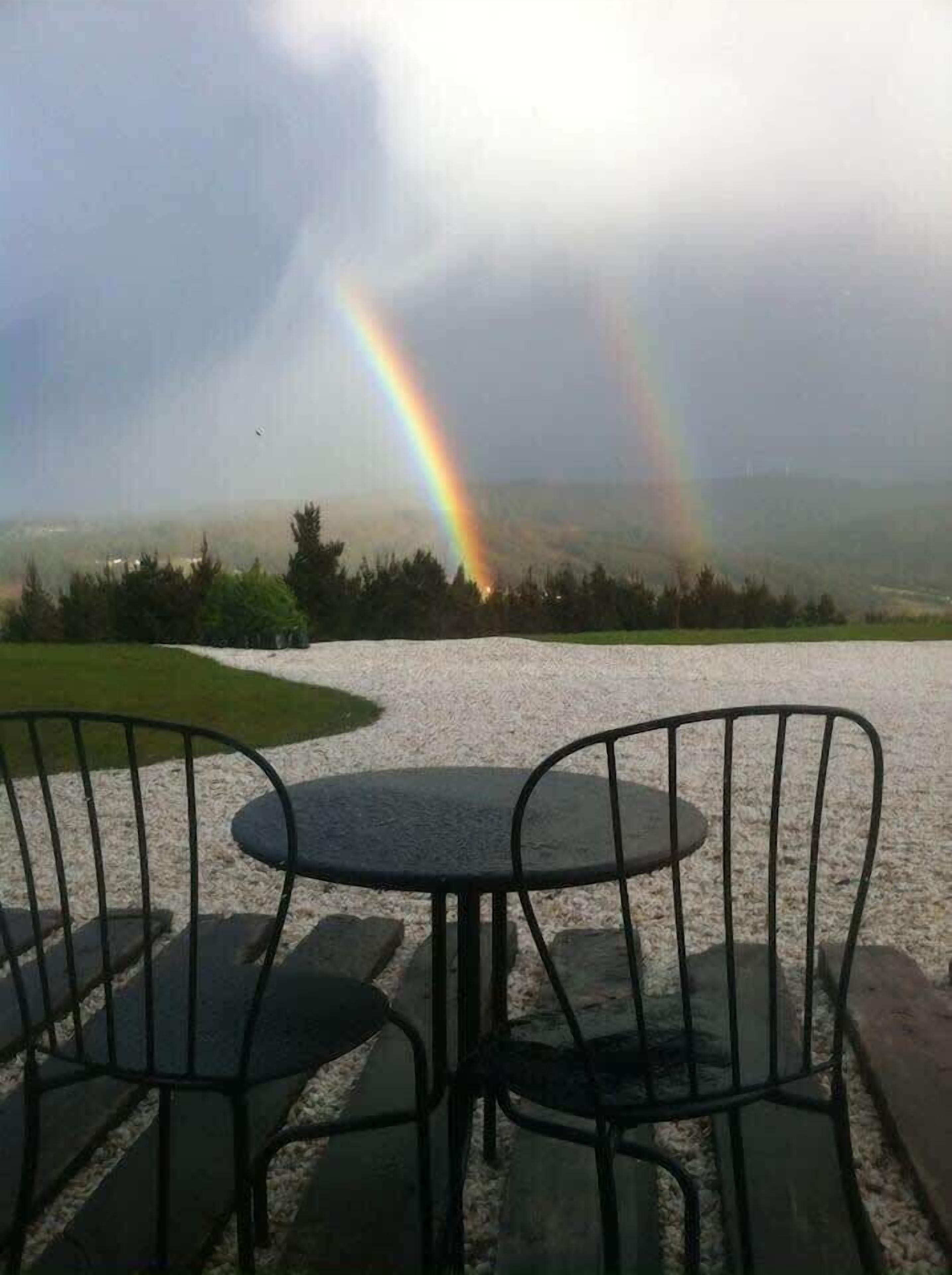 Une table et des chaises sont disposées à l’extérieur, offrant une vue sur un paysage et un arc-en-ciel au loin.