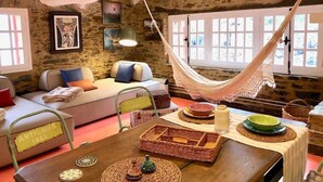 A rustic interior with a hammock, stone walls, a sofa, and a dining table with wicker baskets. - The Cliffs - Cala Porto do Val (O Vicedo)