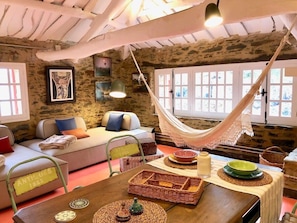 A cozy room with a hammock, rustic stone walls, a sofa, and a dining table with various items.