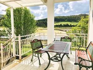 A covered patio with a table and chairs, a white railing, and a view of a green landscape. - The Cliffs - Esteiro Saltwater Beach (O Barqueiro)