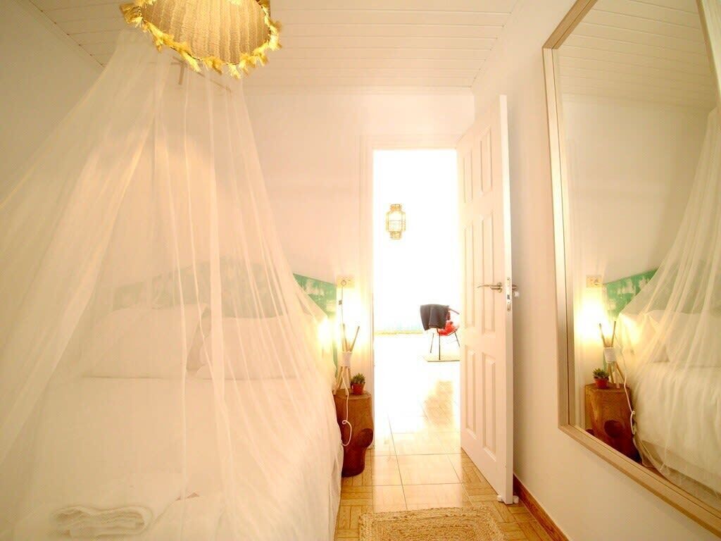 Quarto com uma cama coberta por um dossel branco, um espelho grande na parede e uma porta que dá para outro quarto.