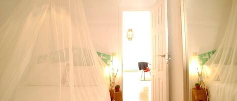 Quarto com uma cama coberta por um dossel branco, um espelho grande na parede e uma porta que dá para outro quarto.