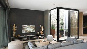 A modern living room with a grey sofa, a round ottoman, a bookshelf, and a large window with a view of a stone wall and plants.