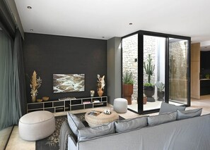 Un salón moderno con un sofá gris, un otoman redondo, una estantería y un amplio ventanal con vistas a una pared de piedra y plantas.