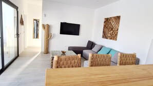 A modern living room with a glass door leading to a balcony, a large cactus, a flat-screen TV, and a wooden wall art piece.