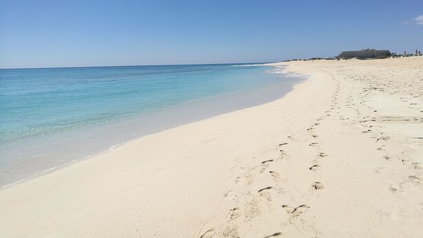 A wide sandy beach with clear blue water and a few footprints.