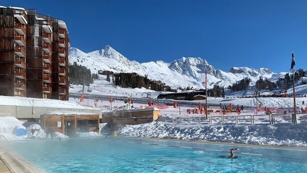 Ein Schwimmbecken mit klarem Wasser, umgeben von verschneiten Bergen und einem Skiort im Hintergrund.