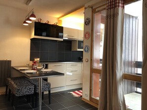 Eine kleine Küche mit schwarzen Fliesen, einem Esstisch mit Stühlen und einem Fenster mit Vorhängen.