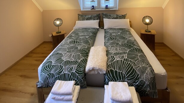 A bedroom with a sloped ceiling, two bedside tables with lamps, a bed with patterned bedding, and a window with blinds. - Beautiful three room apartment. Bright, stylish, cozy (Sachsen - Borna)